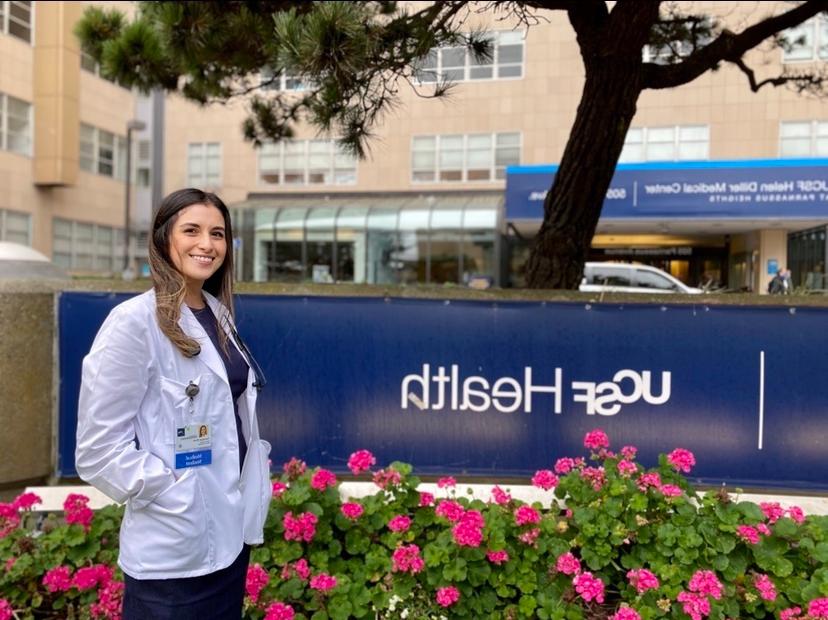 A young woman with long hair in a white coat stands next a UCSF 健康 sign outside, smiling