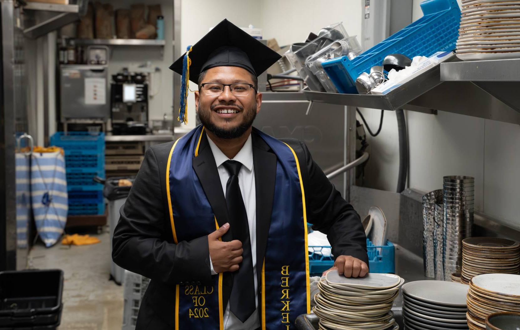 A man with a beard smiles wearing his cap, gown and stole in a commercial kitchen