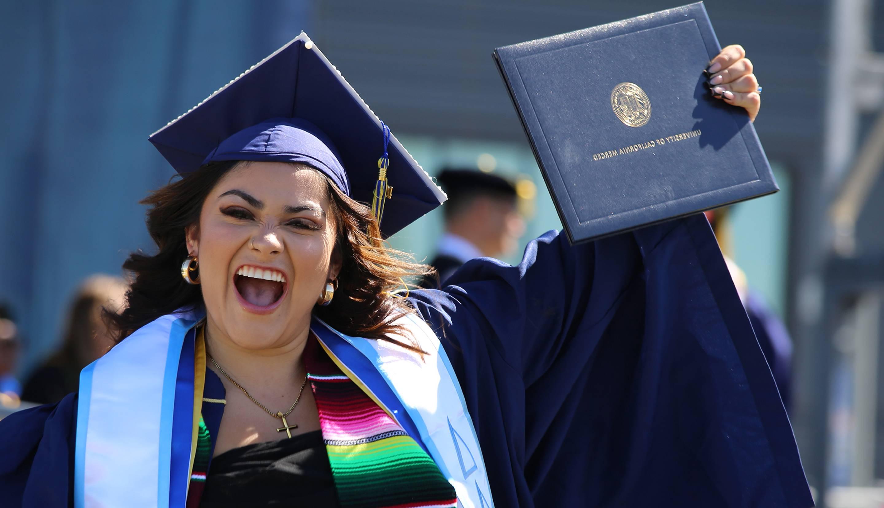 A happy woman graduate holds up her diploma and bears a huge grin as she crosses the stage in cap and gown